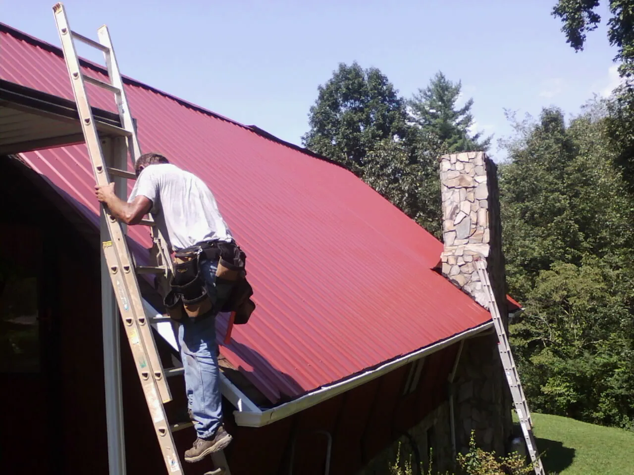 Roofer on a ladder beside a red metal roof and stone chimney — Soffit & Fascia Repair by Roofing Brothers in Kirklin